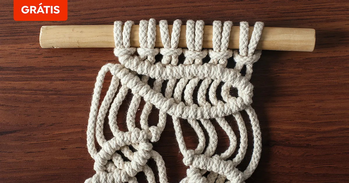 Mãos passando um cordão branco por uma peça de madeira durante a confecção de um suporte de macramê.